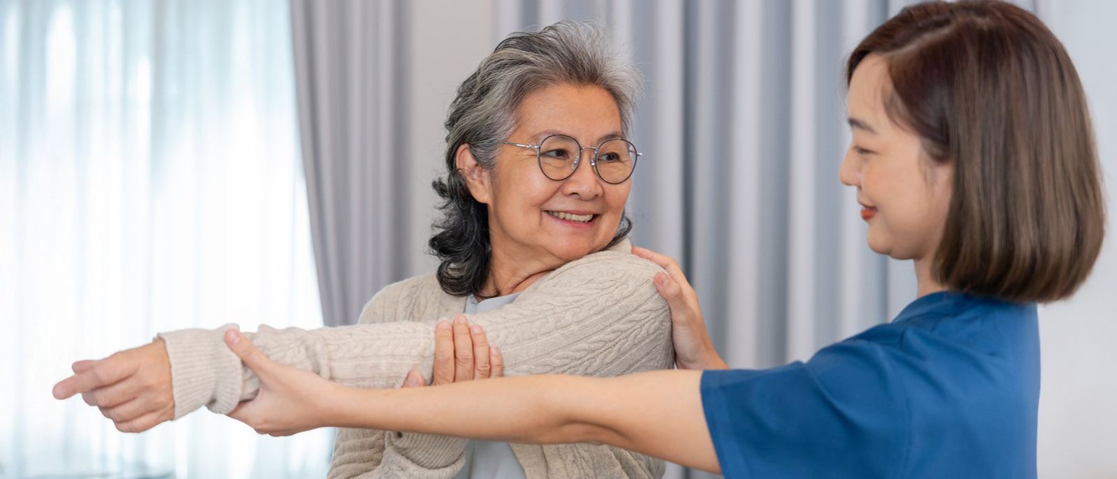 Home health therapist guiding an older woman through an arm stretching exercise in her living room