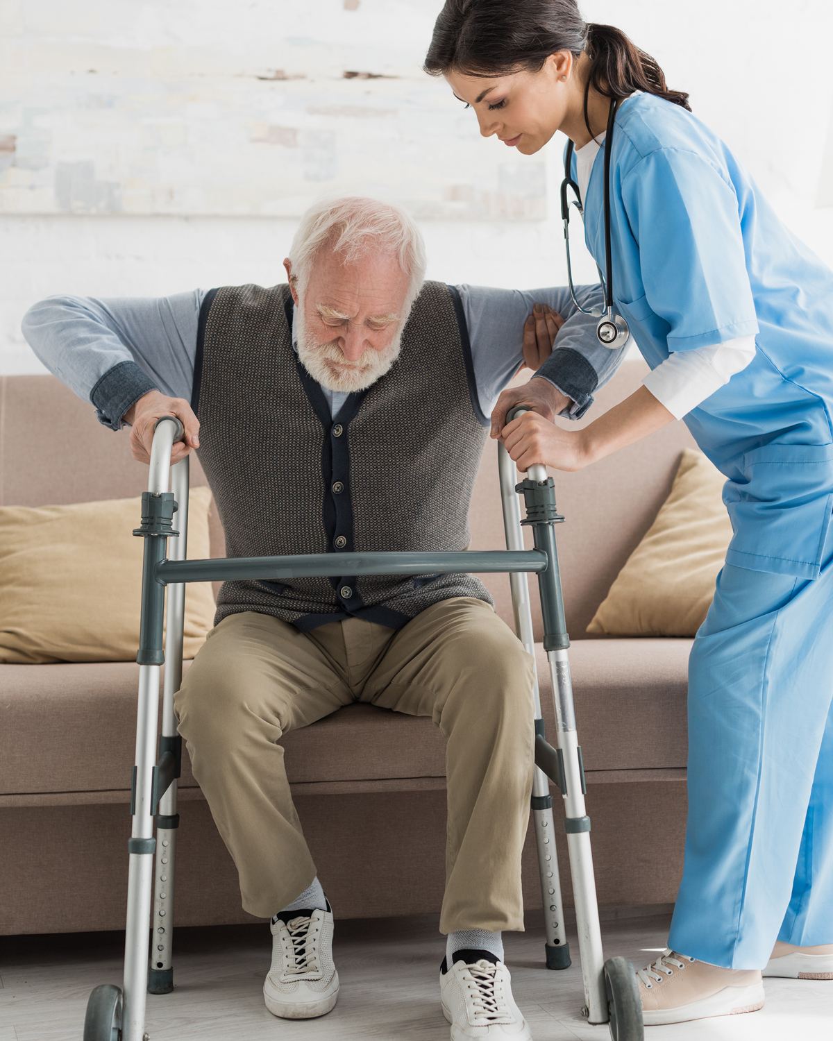 Nurse steadying an older man as he rises from his sofa with a walker at home after surgery
